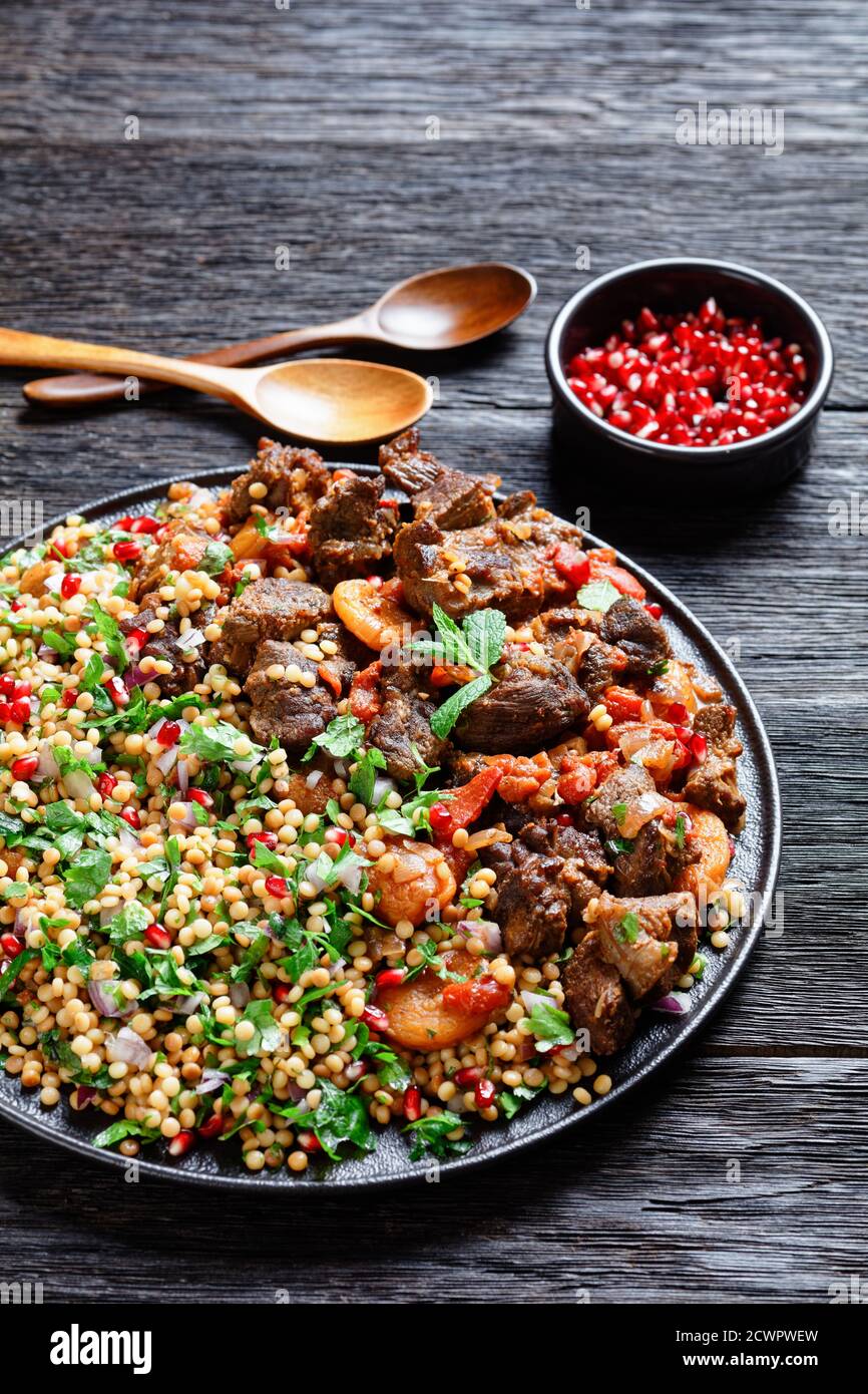Moroccan Lamb Stew with Herbed Pearl Couscous and pomegranate seeds on a black plate on a dark wooden table, vertical view from above Stock Photo