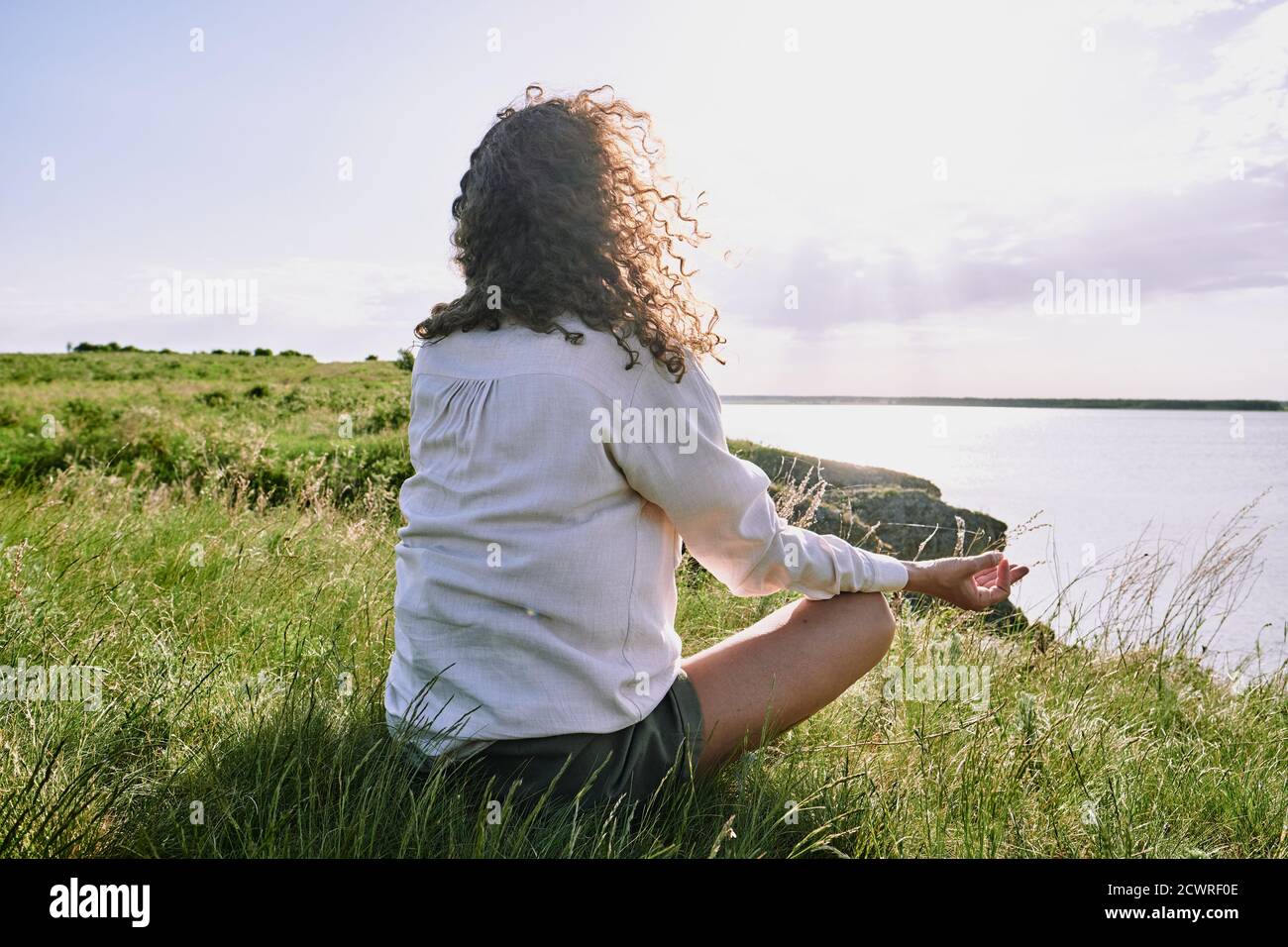 Rear view of curly-haired woman sitting with crossed legs on grass and meditating in silence by tranquil lake Stock Photo