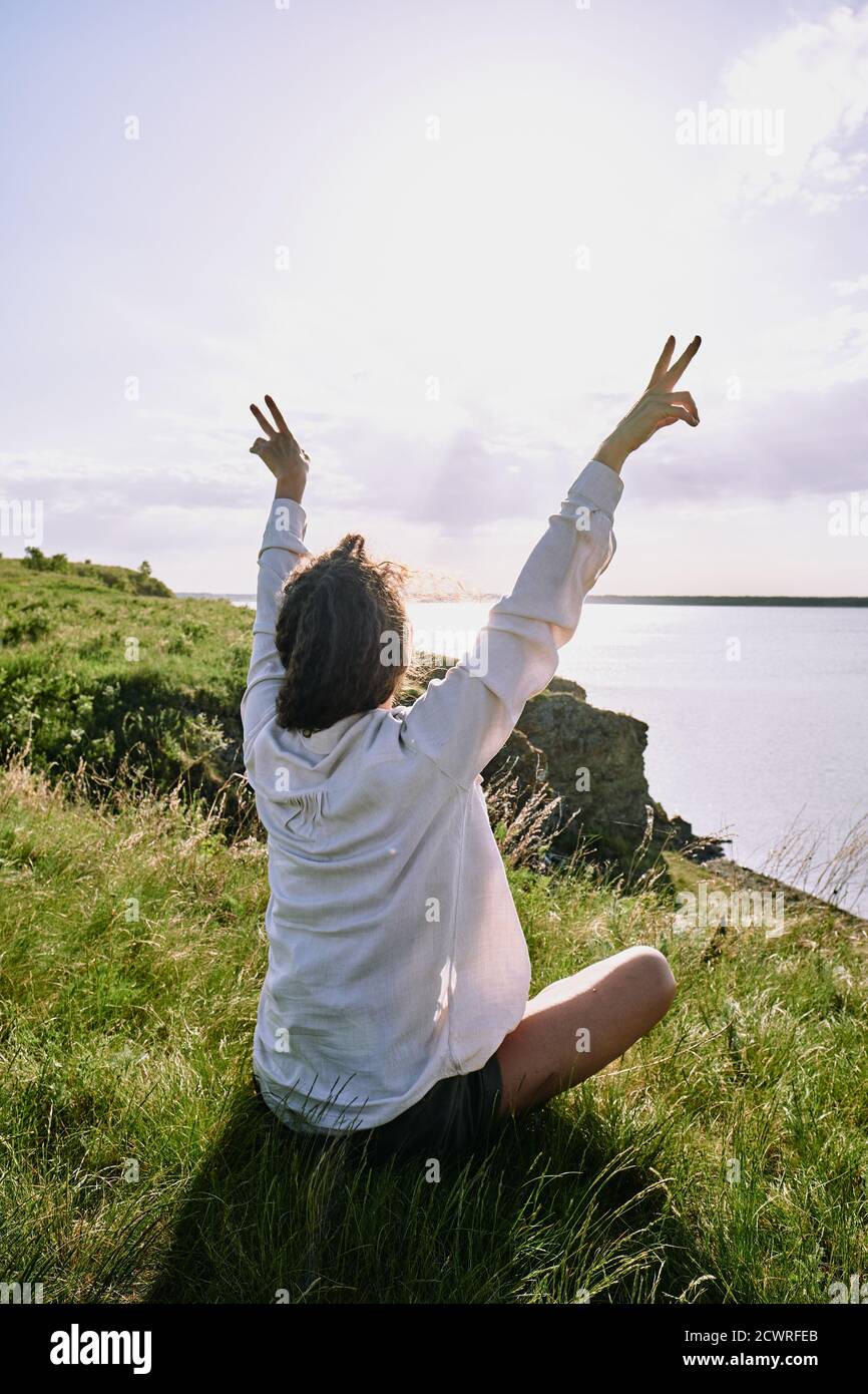 Rear view of young woman in white casual shirt sitting with crossed legs on grass and showing peace signs while contemplating nature Stock Photo