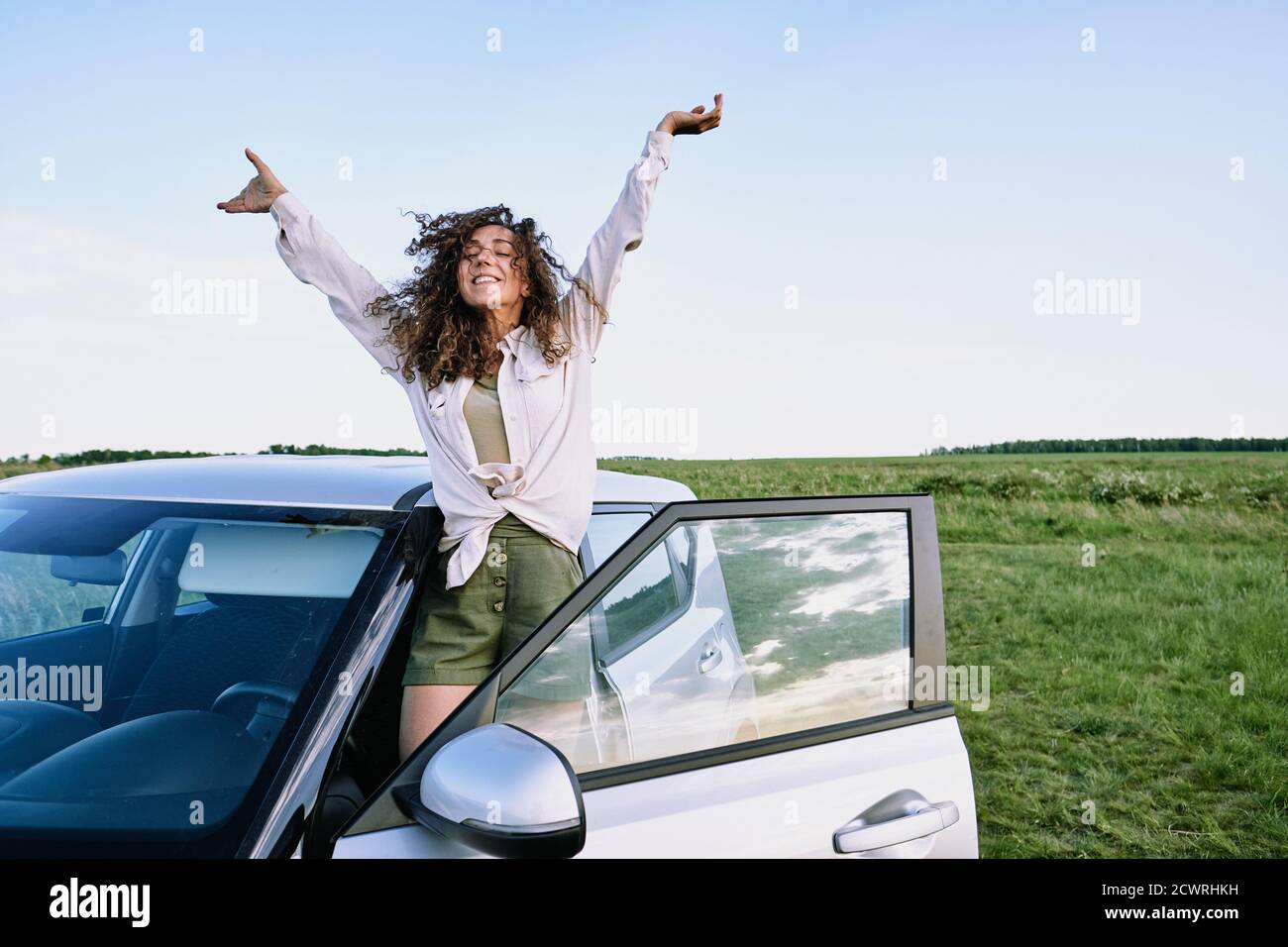 Ecstatic curly-haired girl in casual shirt raising arms and standing on car sill while having fun during car trip Stock Photo