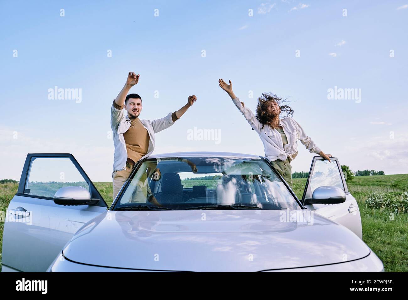 Cheerful excited young couple standing on car sills and waving arms