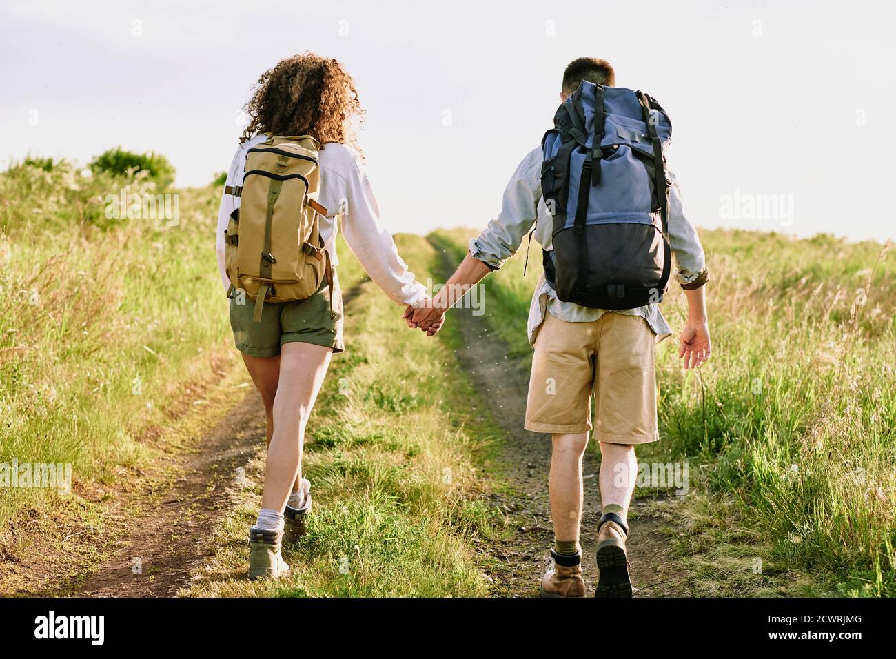 Rear view of young backpackers in shorts holding hands and walking over country road Stock Photo