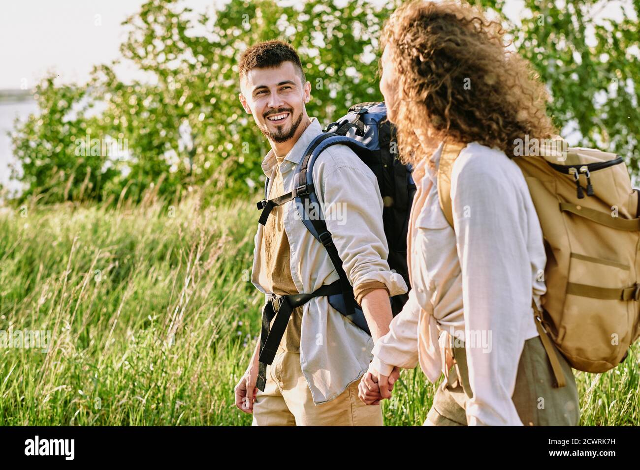 Positive young couple with backpacks holding hands and looking at each other while enjoying hike in country Stock Photo