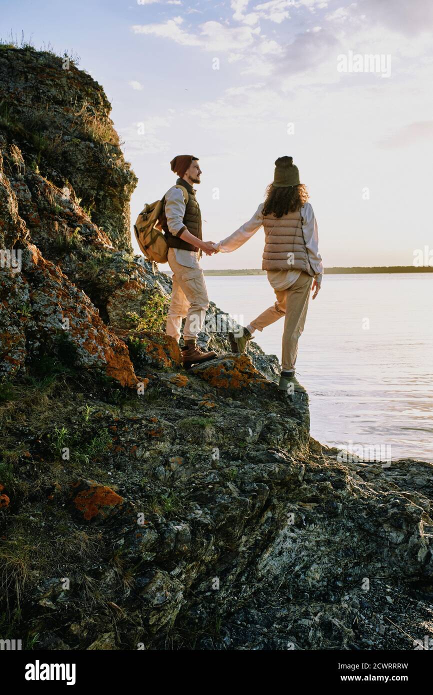 Affectionate hikers in casual clothes standing on rocks of cliff and looking at lake while walking together at sunset Stock Photo