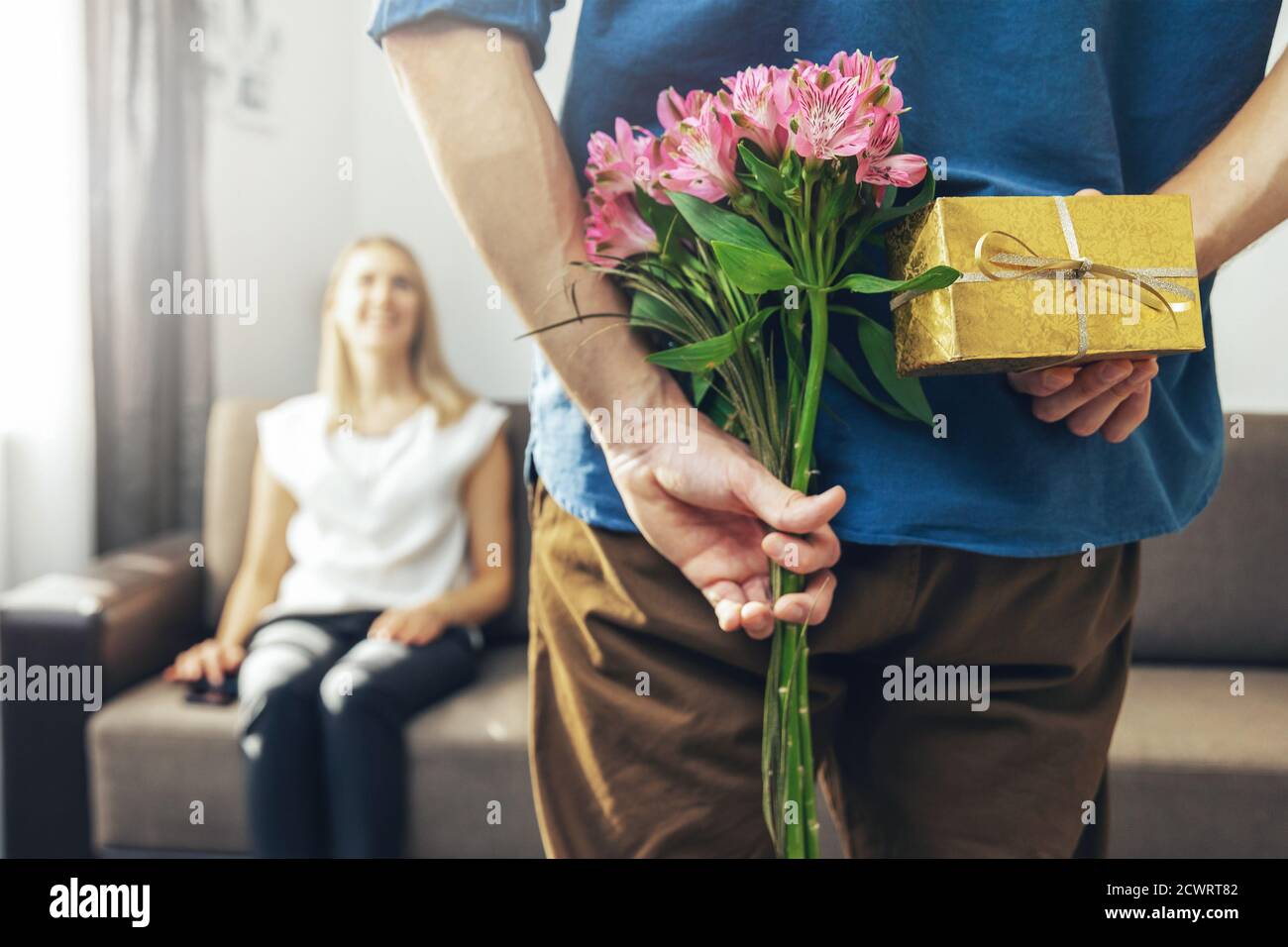 husband hiding romantic surprise present and flowers behind back to beloved wife at home Stock Photo