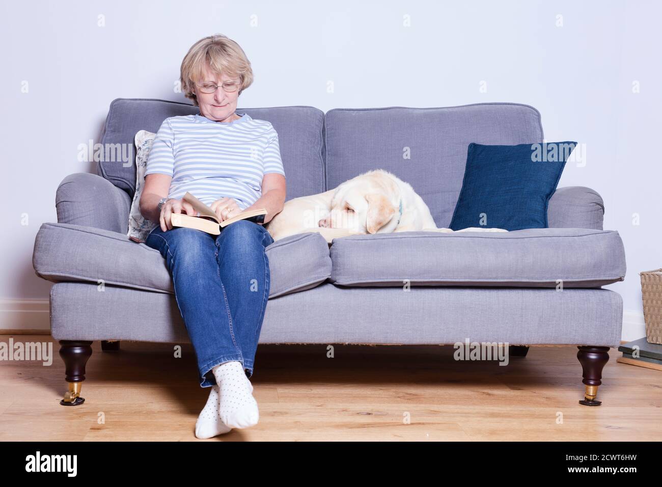 Lady in her late 60s relaxing on the sofa with a book and her dog Stock ...