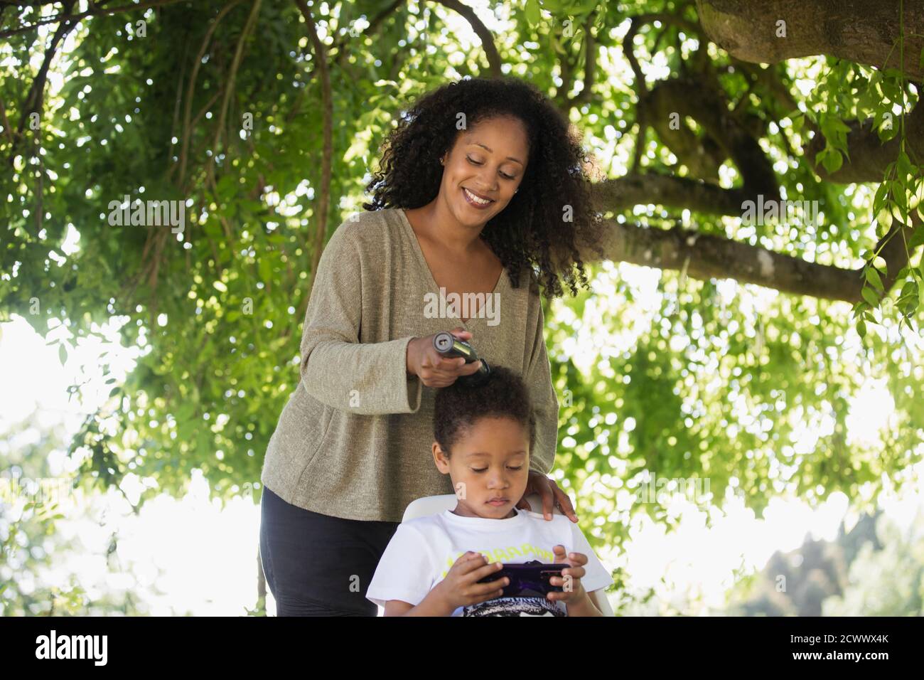 Mother trimming hair of son under tree in summer backyard Stock Photo