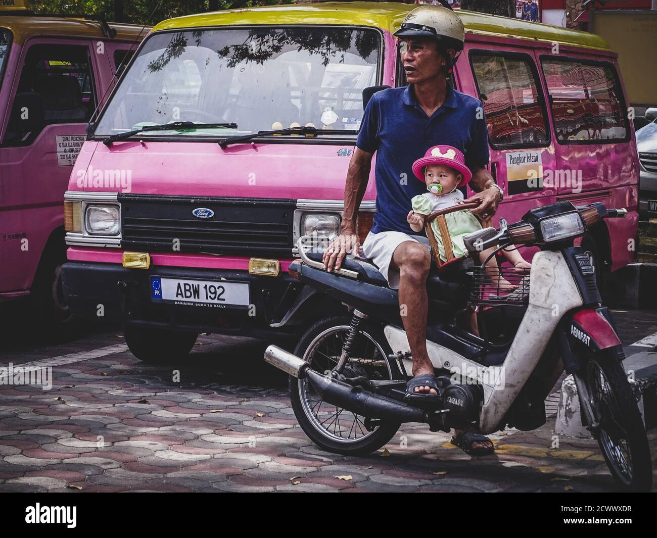 An Asian man with his daughter on a motorcycle shot by the colectivo pick-up spot on Pangkor Island, Malaysia Stock Photo