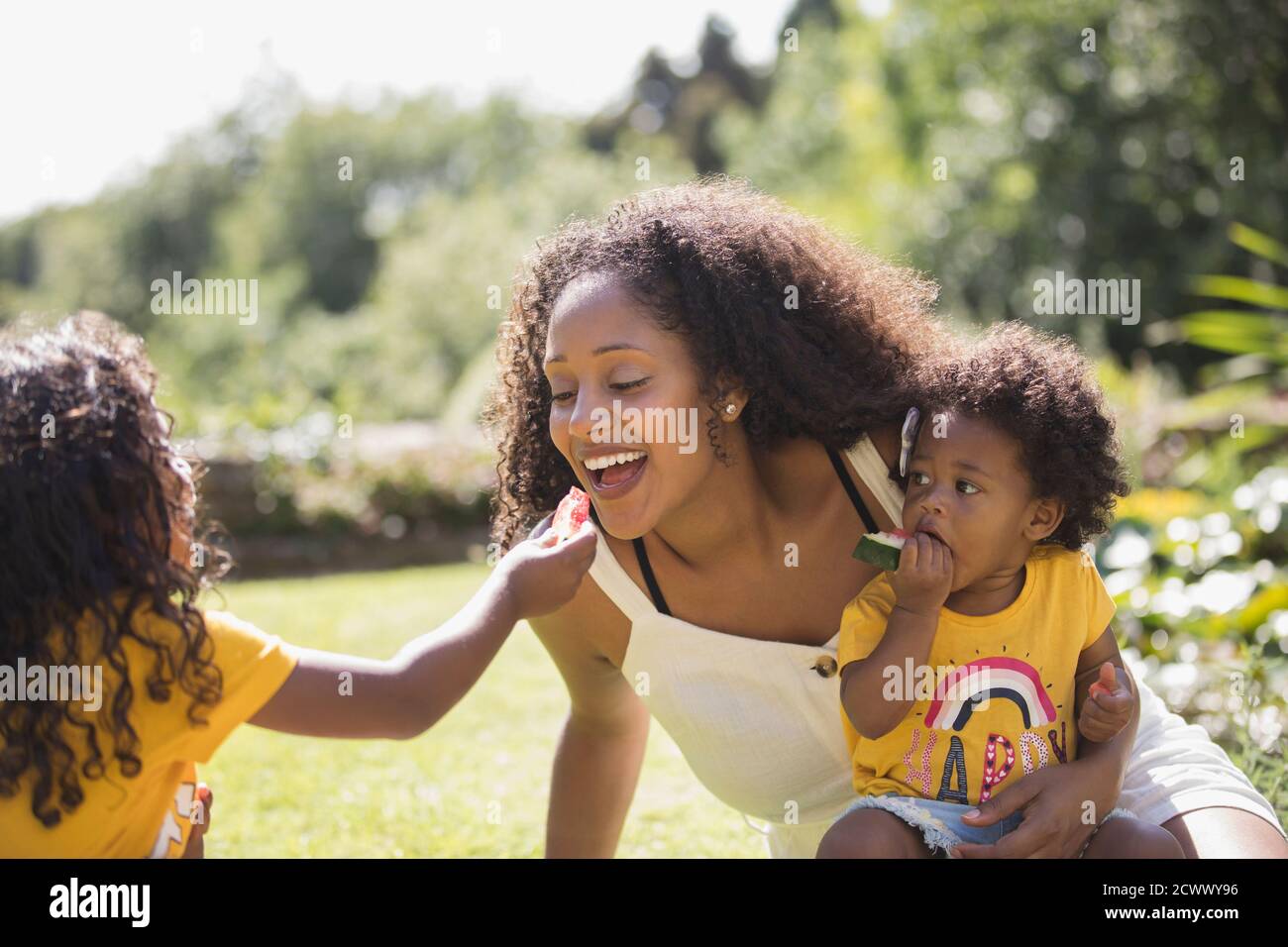 Daughter feeding watermelon to happy mother in sunny backyard Stock Photo