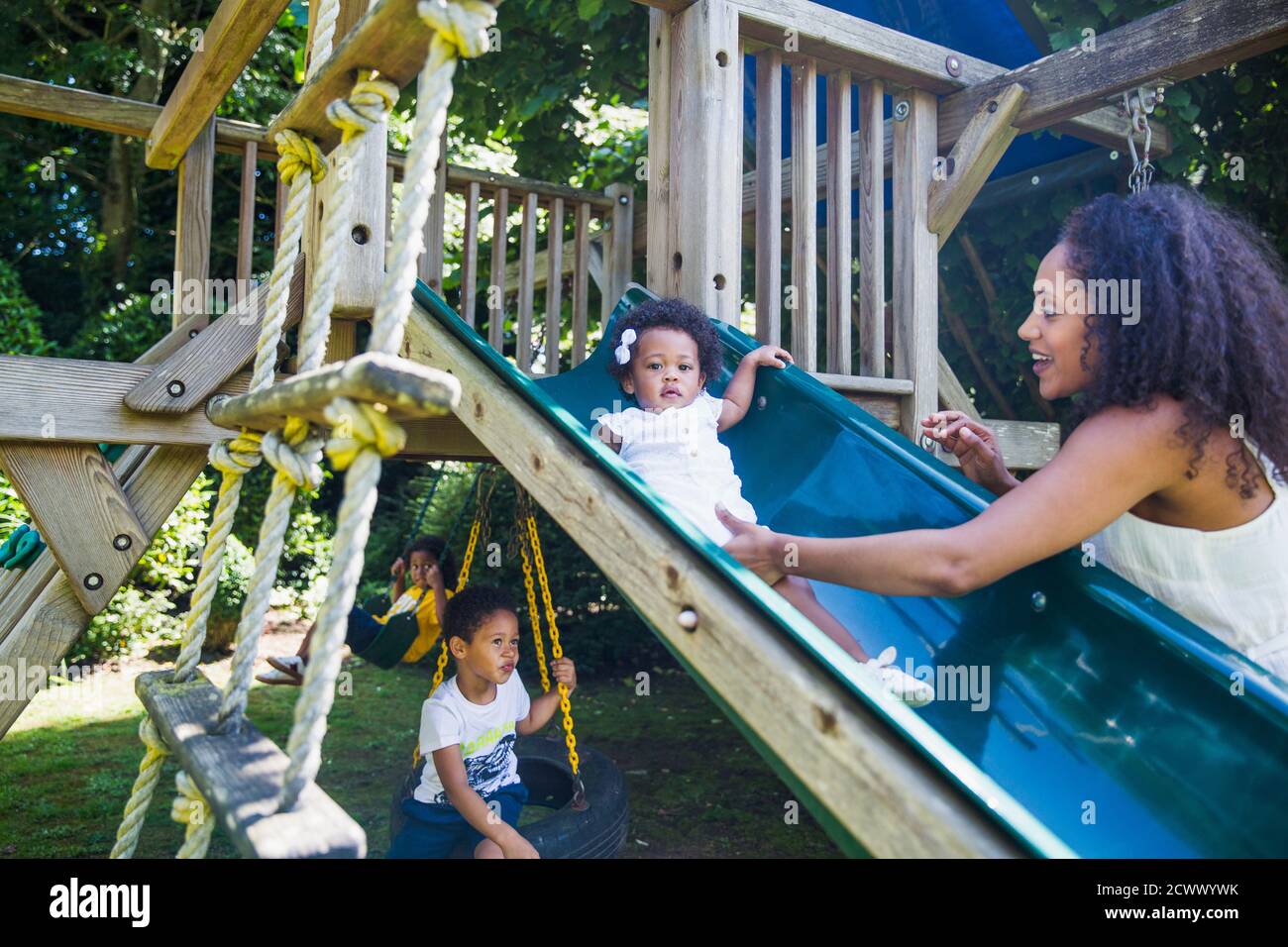 Mother watching cute toddler daughter on playground slide in backyard Stock Photo
