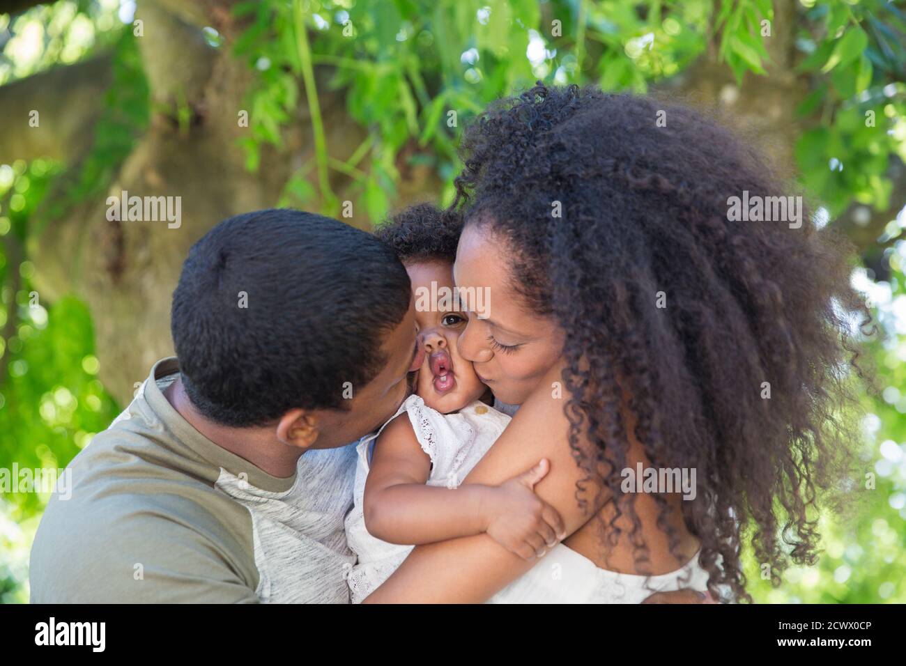 Affectionate parents kissing toddler daughter below tree Stock Photo