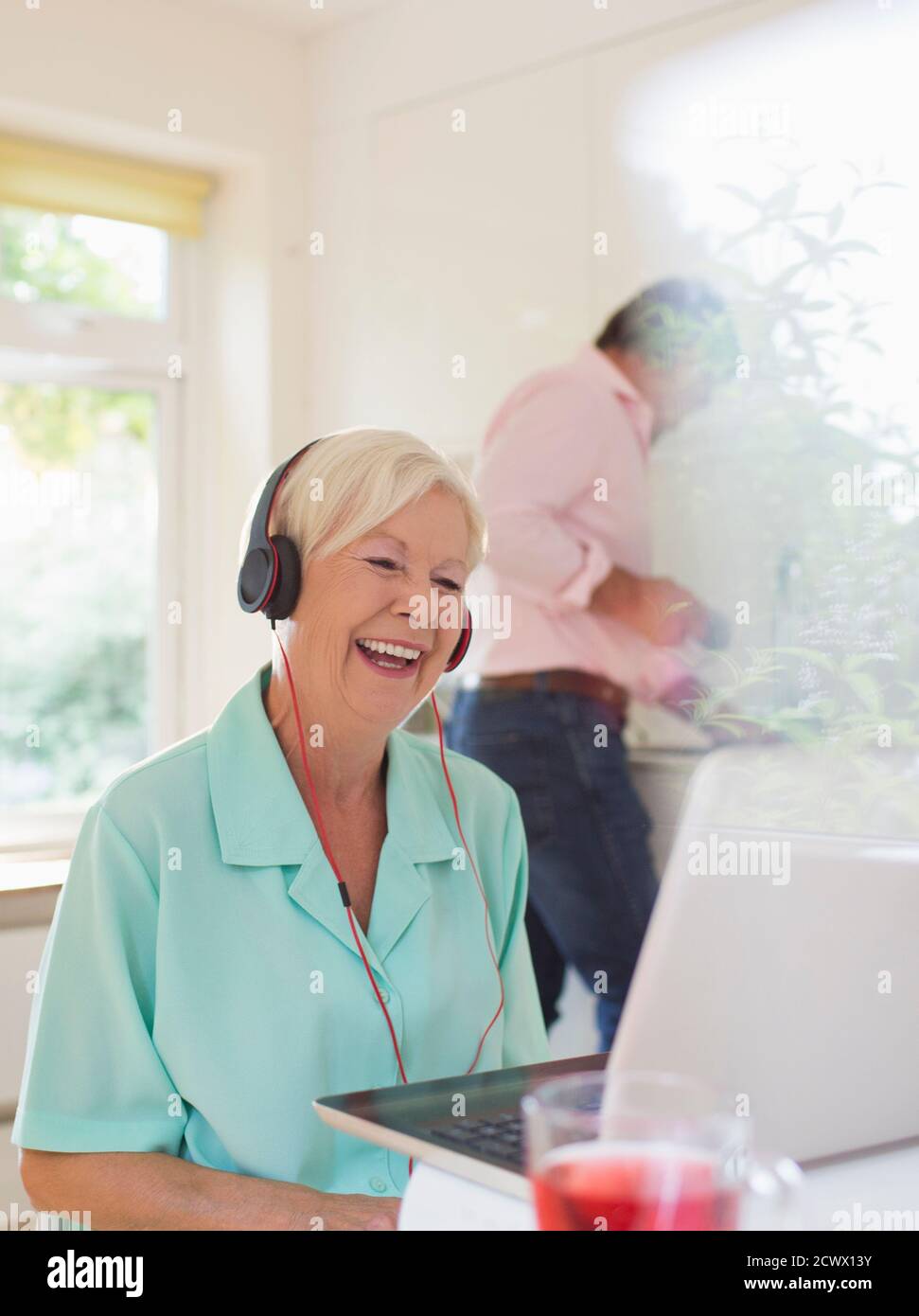 Happy senior woman with headphones video chatting at laptop in kitchen Stock Photo