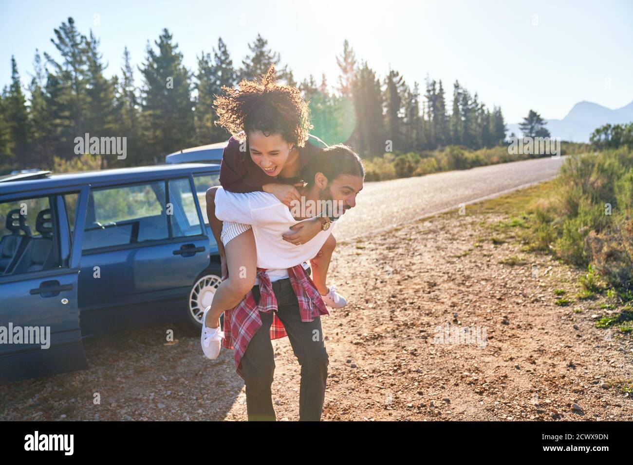 Playful young couple piggybacking outside car at sunny remote roadside Stock Photo