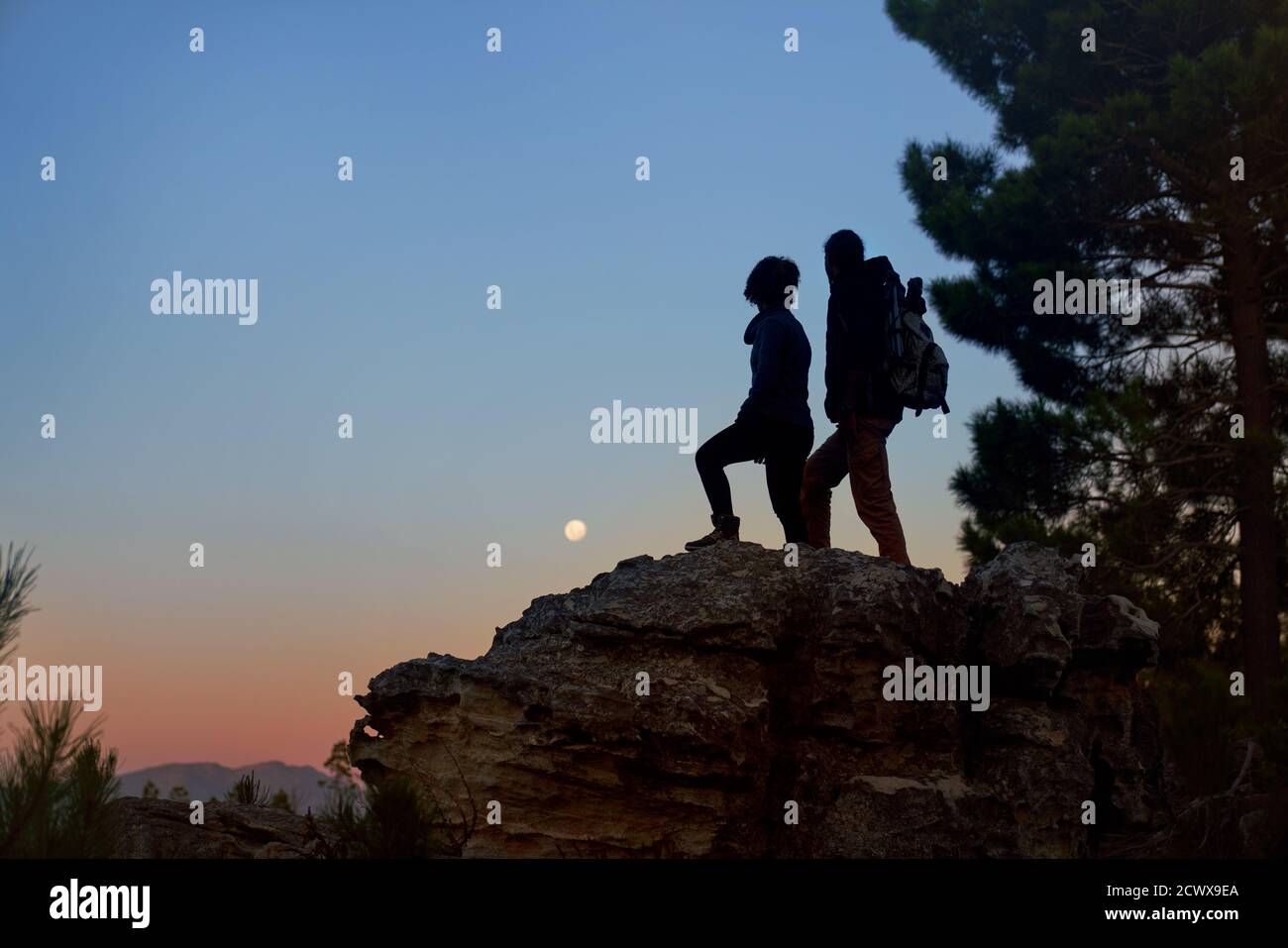 Silhouette young hiker couple enjoying view of moon from rock at dusk Stock Photo