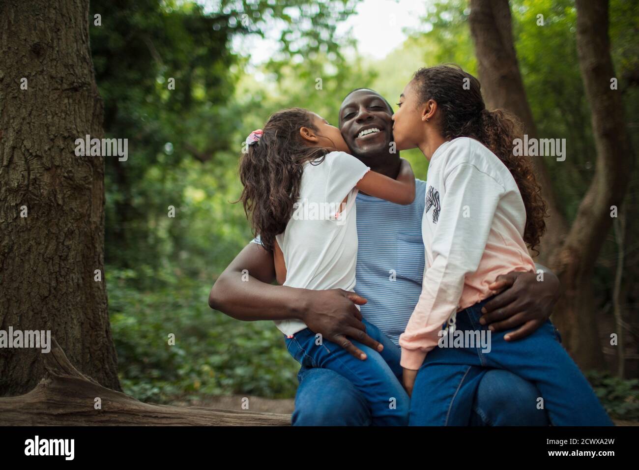 Happy affectionate daughters kissing father below trees in woods Stock Photo
