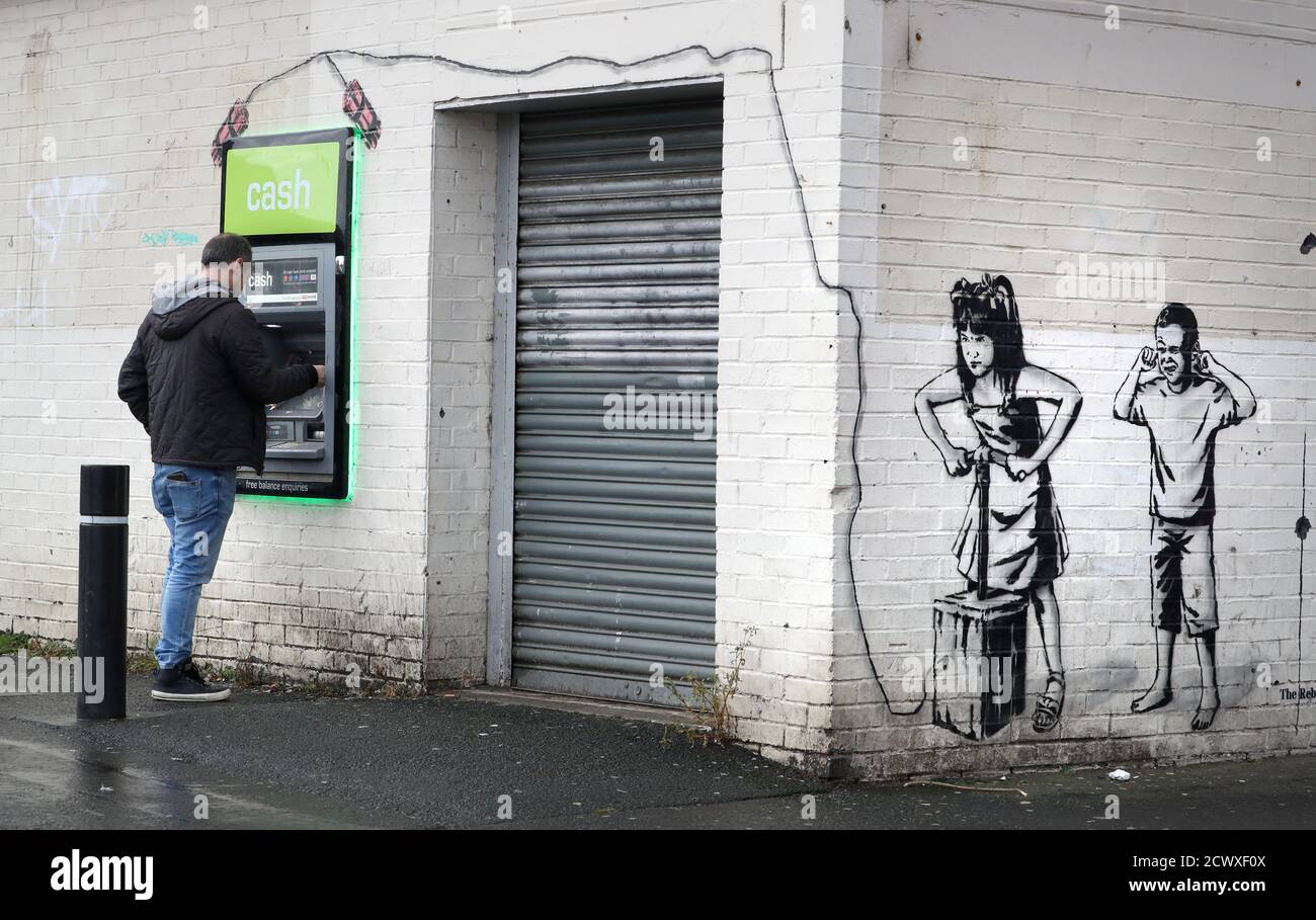 A man uses a cashline next to art work by graffiti artist The Rebel Bear which has recently appeared showing two children about to detonate explosives at the cashline at a shopping centre in Cambuslang. Over recent months the artist has drawn several coronavirus related drawings in Glasgow. Stock Photo