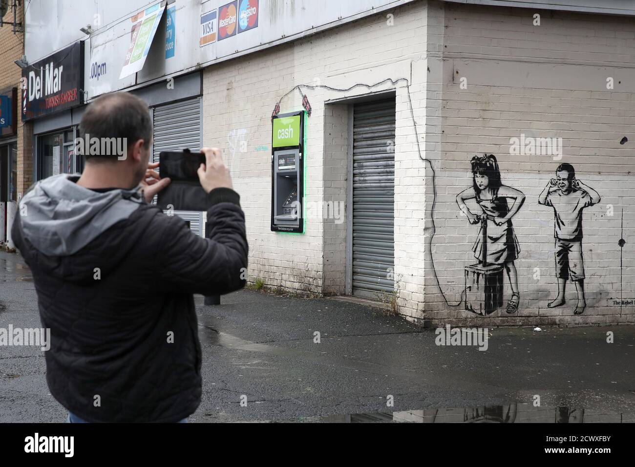A man takes a picture of the art work by graffiti artist The Rebel Bear which has recently appeared showing two children about to detonate explosives at the cashline at a shopping centre in Cambuslang. Over recent months the artist has drawn several coronavirus related drawings in Glasgow. Stock Photo