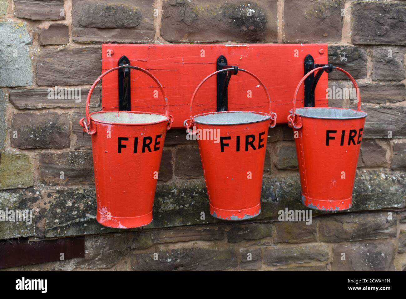 close up of 3 red Fire buckets hanging from hooks on a stone wall outside Stock Photo