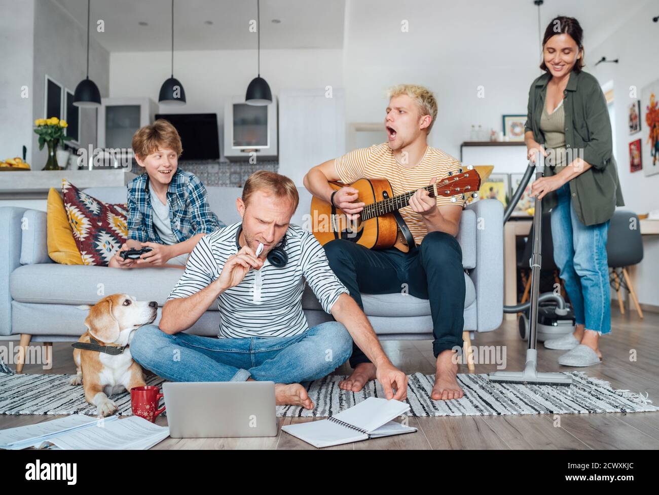 Quarantine time: Family at living room together. Father distance working using laptop, mother cleaning the room, older son playing acoustic guitar and Stock Photo