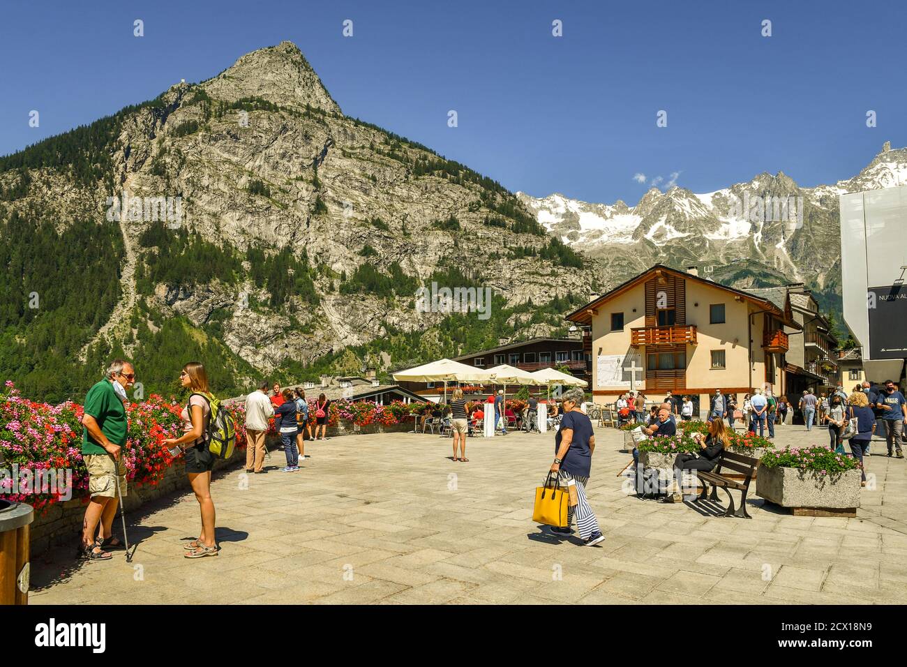 Piazza Abbé Henry, the main square of the famous Alpine town, with tourists and backpackers, Mont Chetif and Mont Blanc massif in summer, Courmayeur Stock Photo