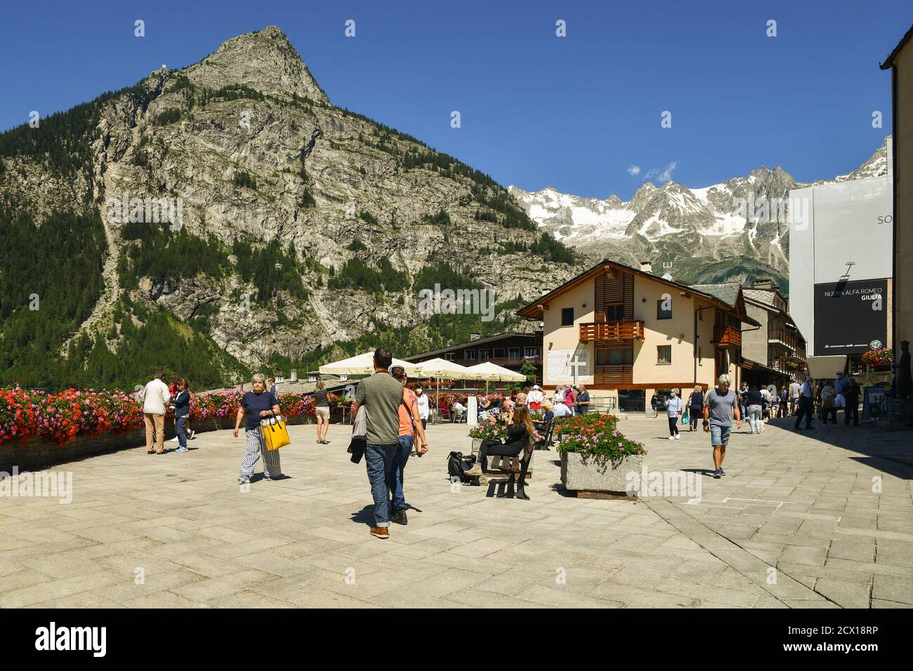 Piazza Abbé Henry, the main square of the famous Alpine town, with tourists and backpackers, Mont Chetif and Mont Blanc massif in summer, Courmayeur Stock Photo