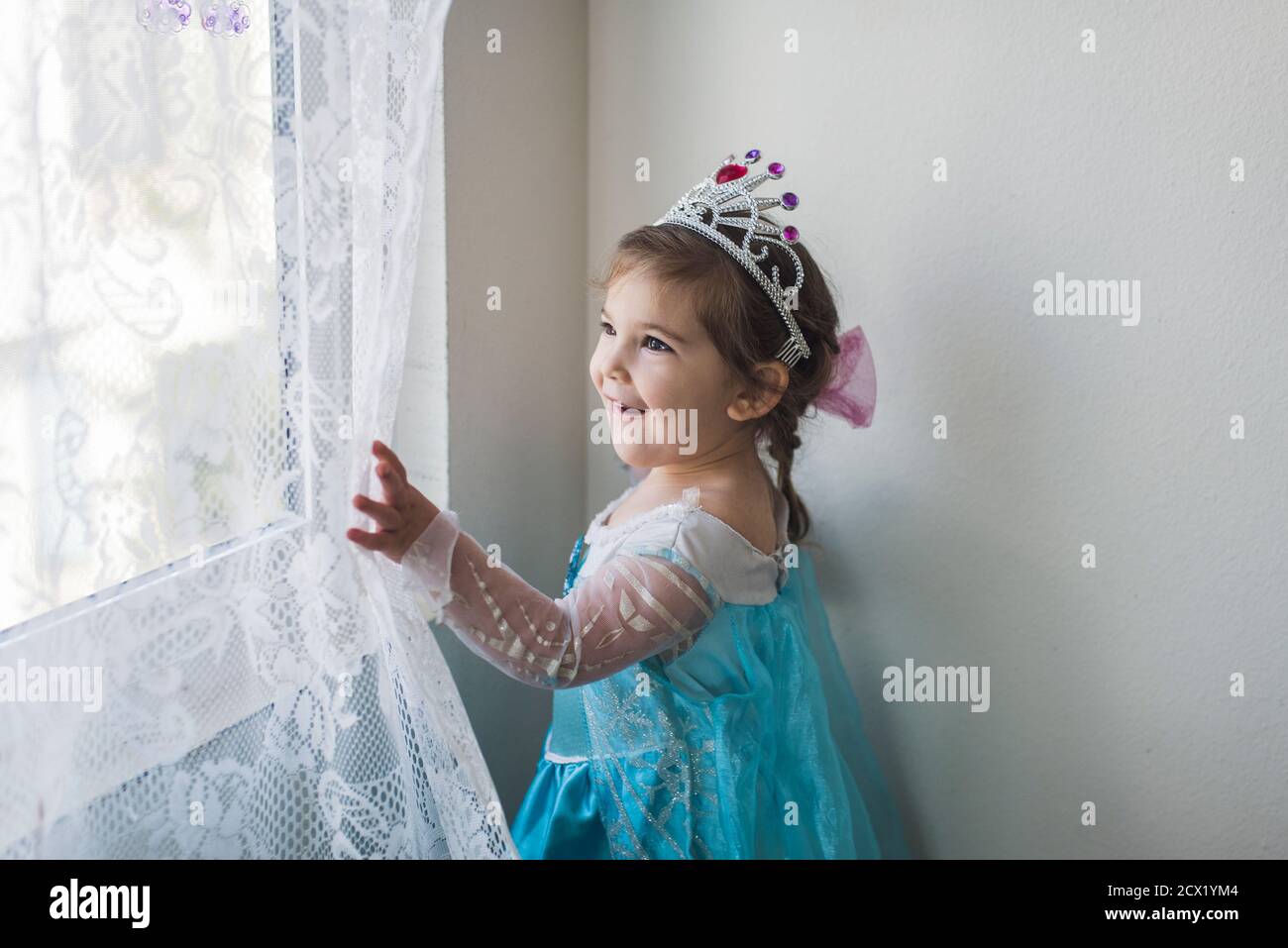 Smiling girl in princess costume and tiara touching white lace curtain Stock Photo