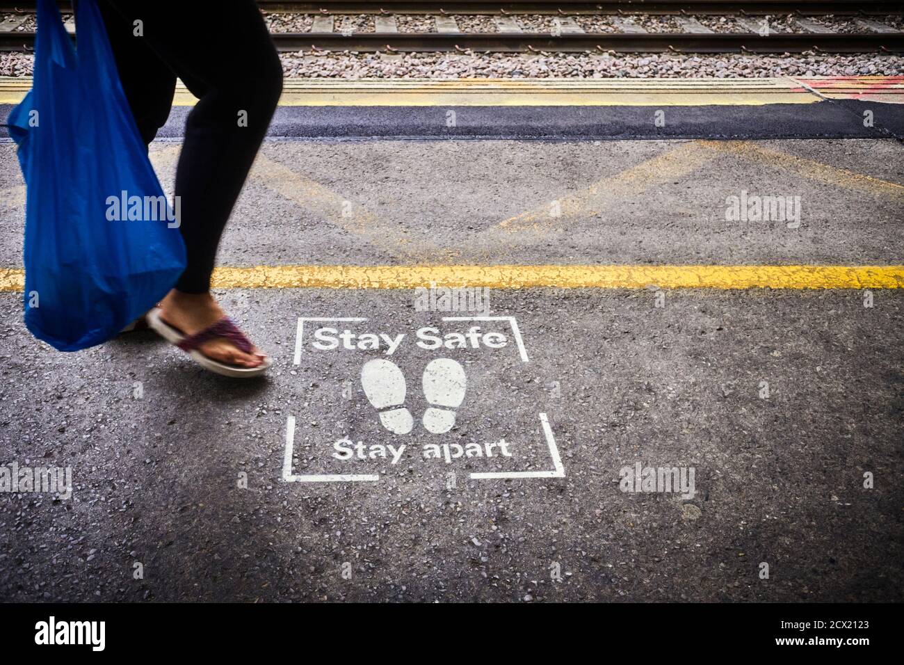 Stay safe, stay apart stencil at the platform edge on Milton Keynes railway station with a woman in open sandels blurred Stock Photo