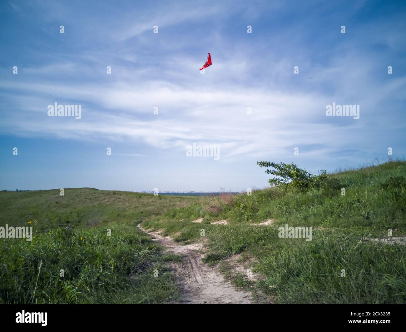 Red delta hang-glider flying high in a beautiful blue sky with white cirrus clouds in Khodosivka, Kyiv. Green hill with brown earthy path and lonely t Stock Photo
