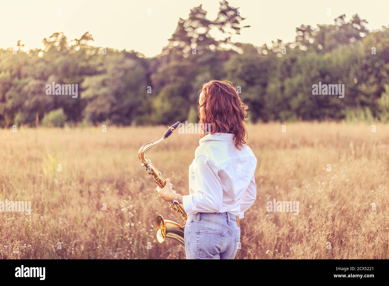 Young red-haired woman in a white shirt with a tenor saxophone in her hands stands in an autumn field and looks at the sunset Stock Photo