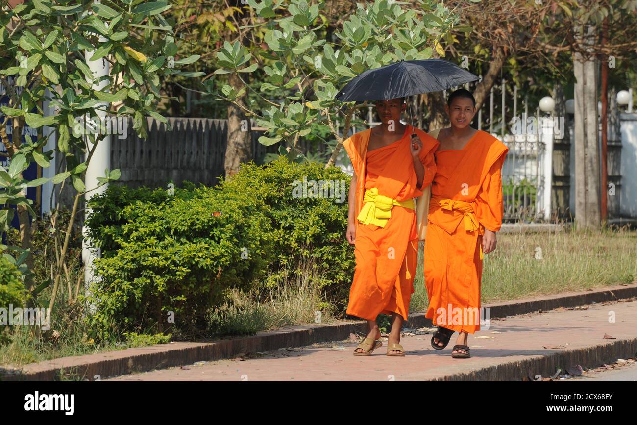 Two monks walking in the street under a parasol. Luang Prabang, Laos.  This image contains culturally relevant material: Each monks robe has three parts, the outer garment called the tricivara the under garment called the uttarasanga and the cloak called the samghati. Stock Photo