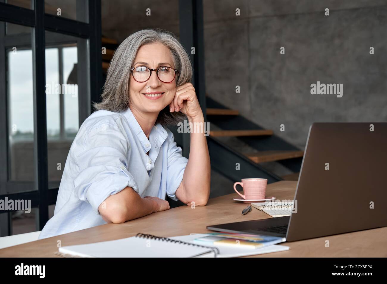 Smiling mature middle aged woman sitting at workplace with laptop, portrait. Stock Photo