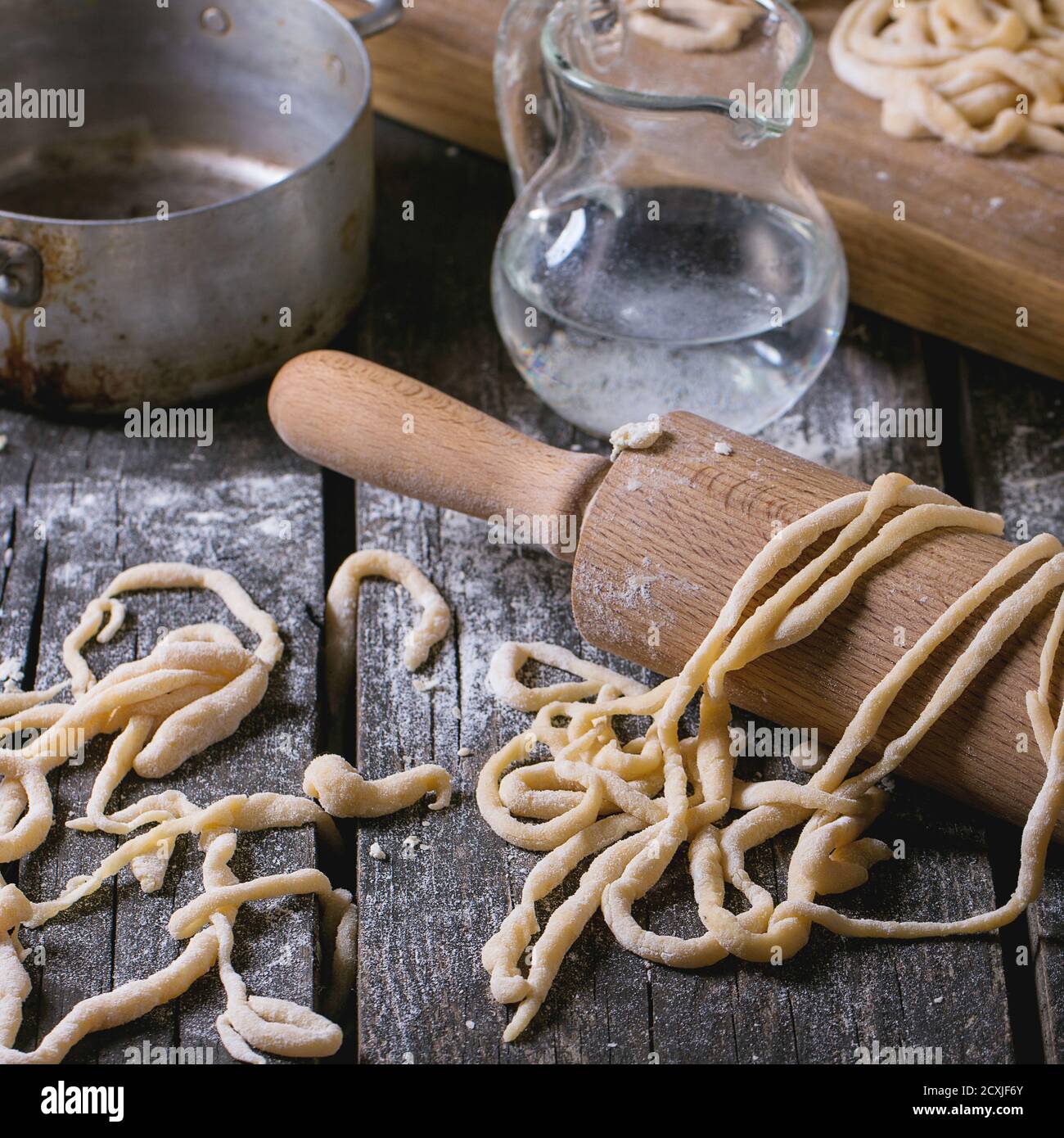 Fresh homemade pici pasta on wood chopping board over old wooden table with flour, aluminum pan, rolling-pin and galss jug of water. Dark rustic style Stock Photo