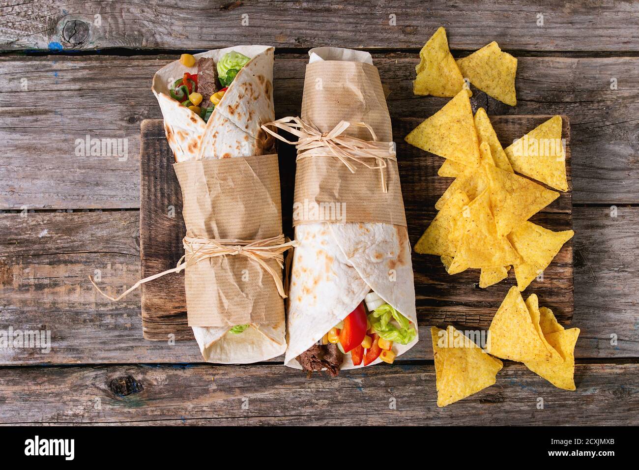 Mexican style dinner. Two papered tortillas burrito with beef and vegetables served with nachos chips over old wooden background. Flat lay Stock Photo