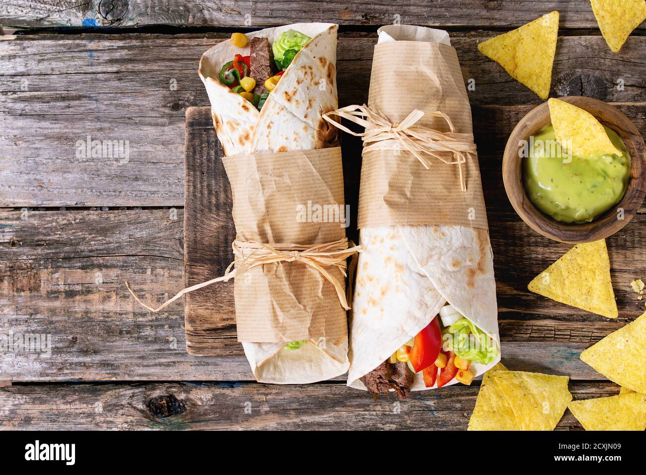 Mexican style dinner. Two papered tortillas burrito with beef and vegetables served with nachos chips and guacomole sauce over old wooden background. Stock Photo