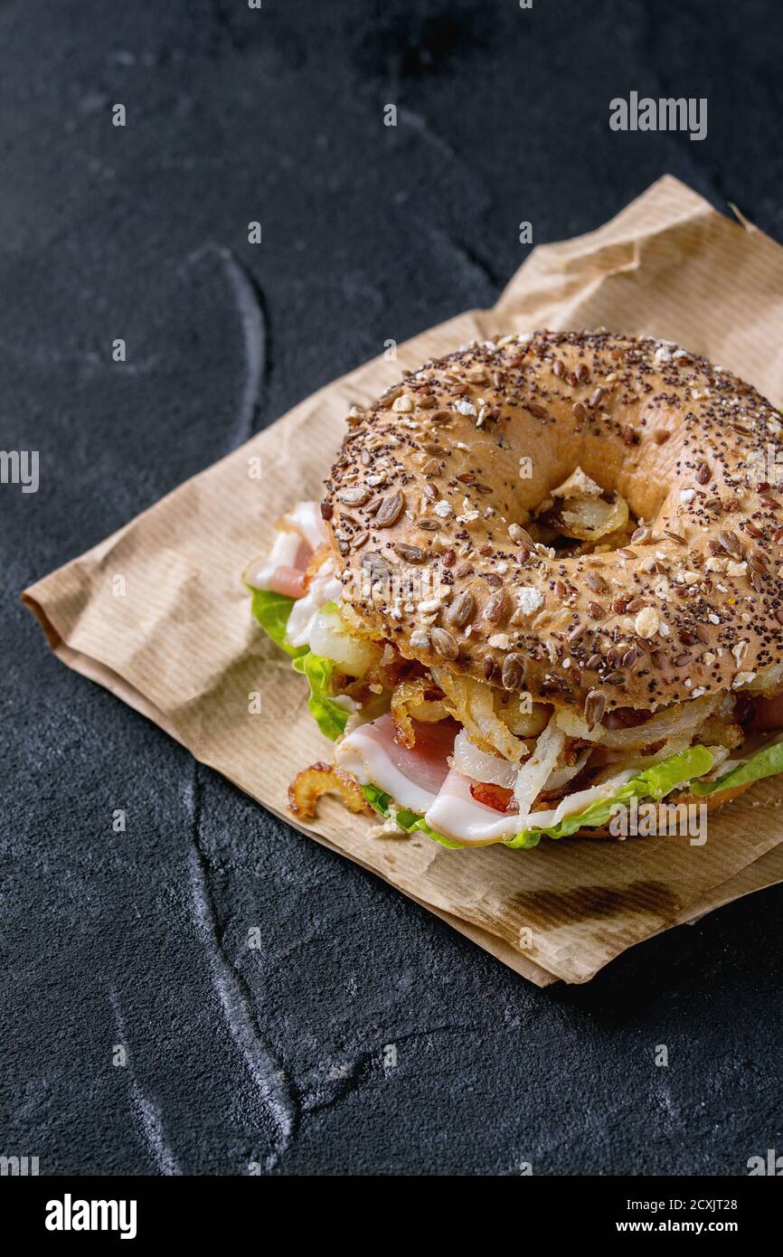Sprinkle seeds Whole Grain bagel with fried onion, green salad and prosciutto ham on paper over black stone textured background. Stock Photo