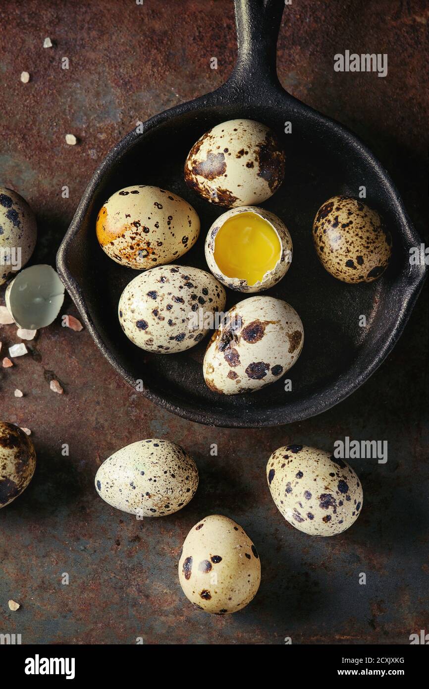 Whole and broken quail eggs with yolk in shell and pink sea salt crystal in small iron cast pan over old rusty texture metal background. Top view with Stock Photo