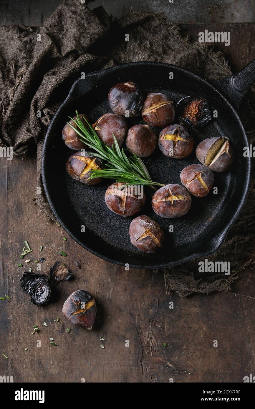 Roasted chestnuts in the ashes with rosemary in cast-iron pan on sackcloth over old dark wooden background. Top view with space for text. Stock Photo