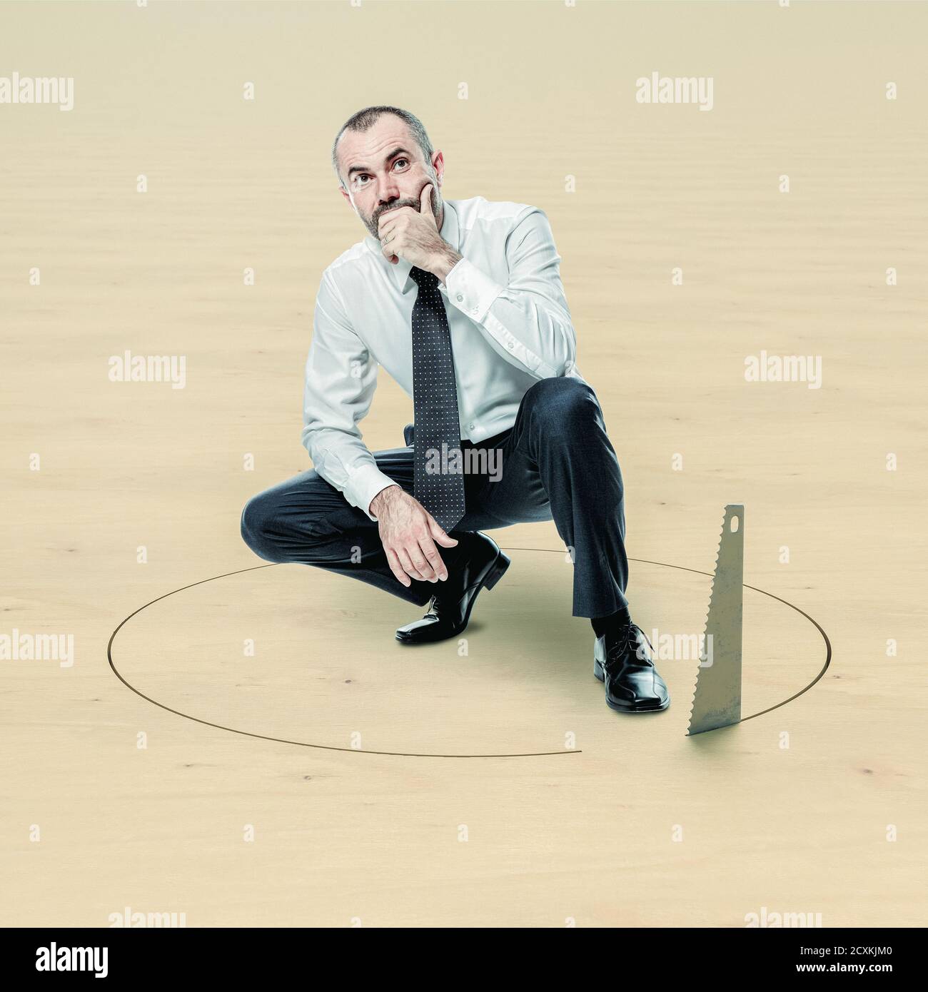 businessman crouched and thoughtful and saw cutting the floor around him creating a hole to make him fall. Stock Photo