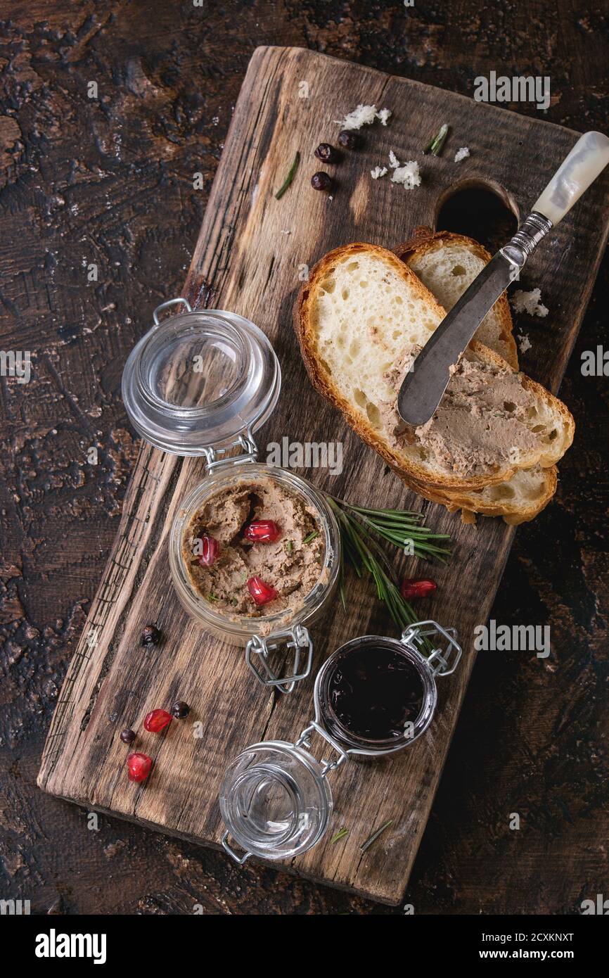 Glass jars of chicken liver pate with blackcurrant jam, pomegranate grain and sliced bread, served with vintage knife on wooden chopping board over da Stock Photo