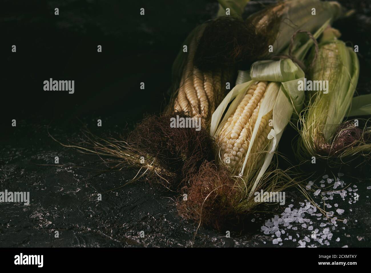 Young raw uncooked corn cobs in leaves and sea salt. Close up over dark brown concrete texture background. Stock Photo