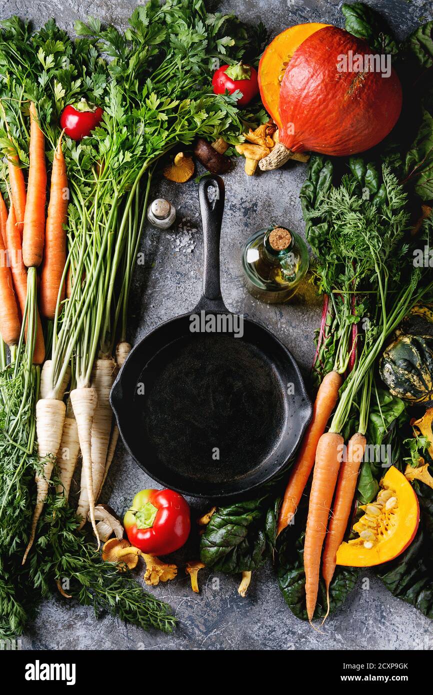 Variety of autumn harvest vegetables carrot, parsnip, chard, paprika, hokkaido pumpkin, mushrooms. Empty iron cast pan, olive oil, salt over gray text Stock Photo