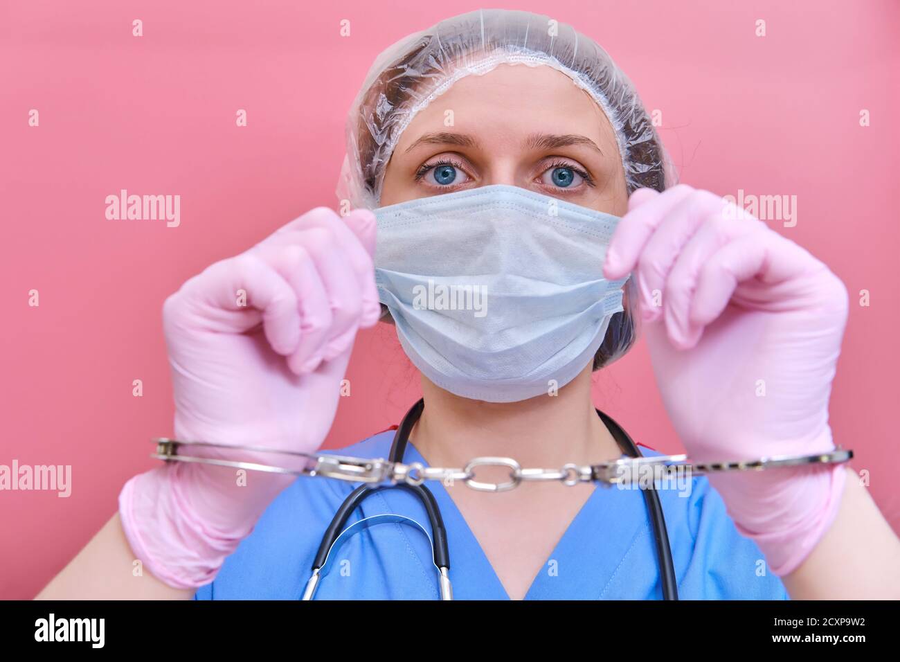 Portrait of a doctor woman in a medical mask in handcuffs on her hands, closeup. Nurse in blue uniform and protective gloves on a pink background. Stock Photo