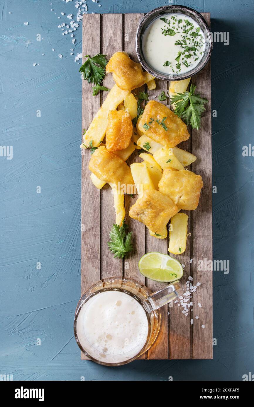 Traditional british fast food fish and chips. Served with white cheese sauce, lime, parsley, glass of lager beer on wooden serving board over blue con Stock Photo