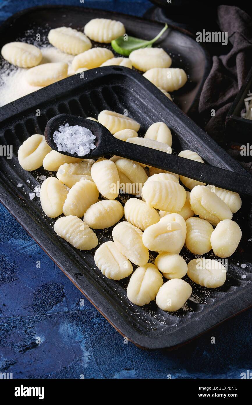Raw uncooked potato gnocchi in black wooden plates with ingredients. Flour, basil, salt over dark blue concrete background. Home cooking. Stock Photo