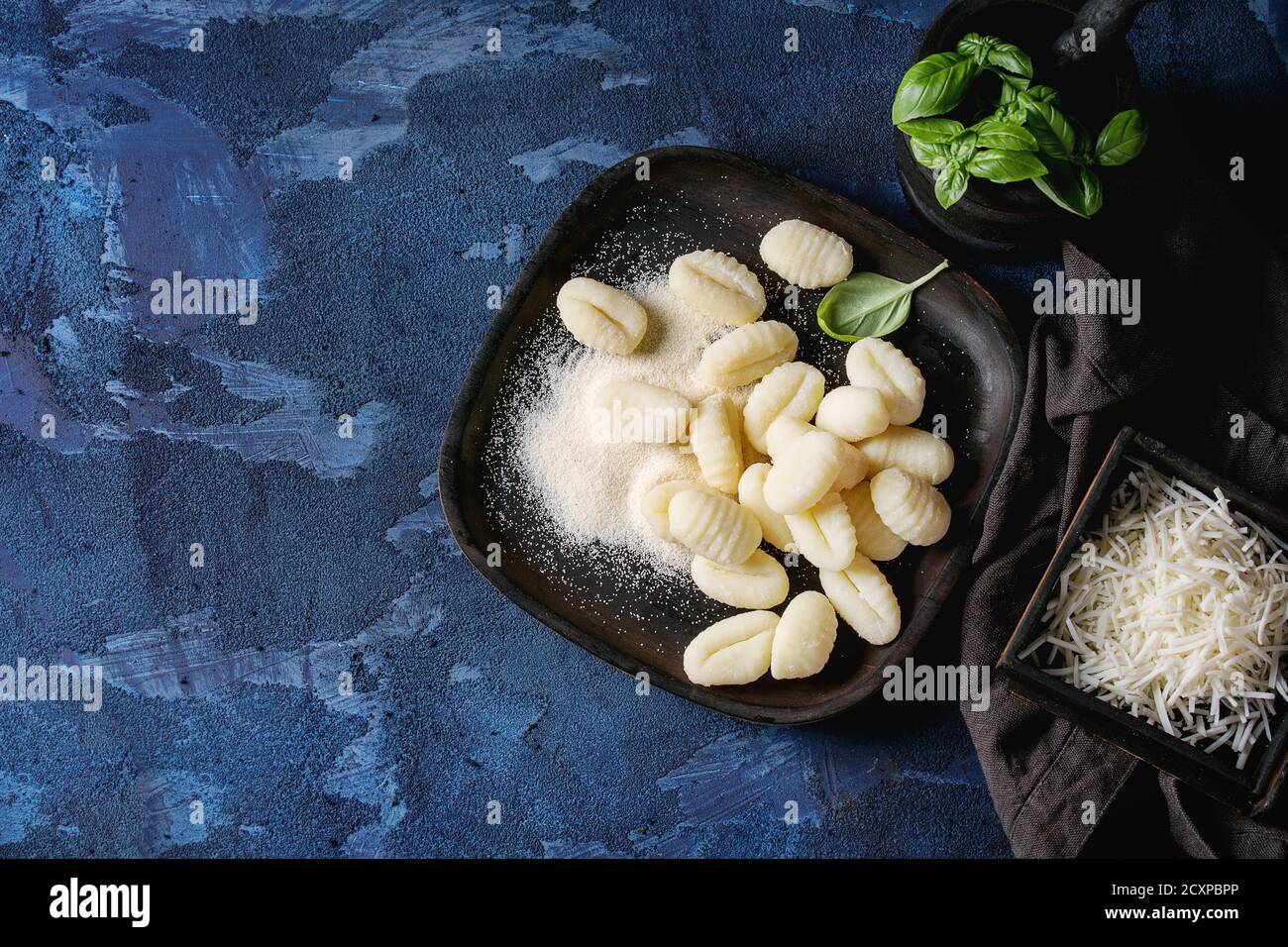 Raw uncooked potato gnocchi in black wooden plates with ingredients. Flour, grated parmesan cheese, basil, over dark blue concrete background. Top vie Stock Photo