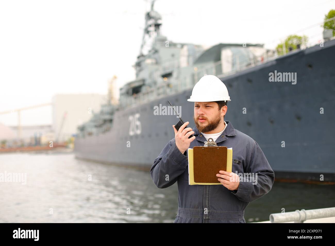 Marine deck officer talking by walkie talkie radio and standing near vessel with documents in hands. Stock Photo