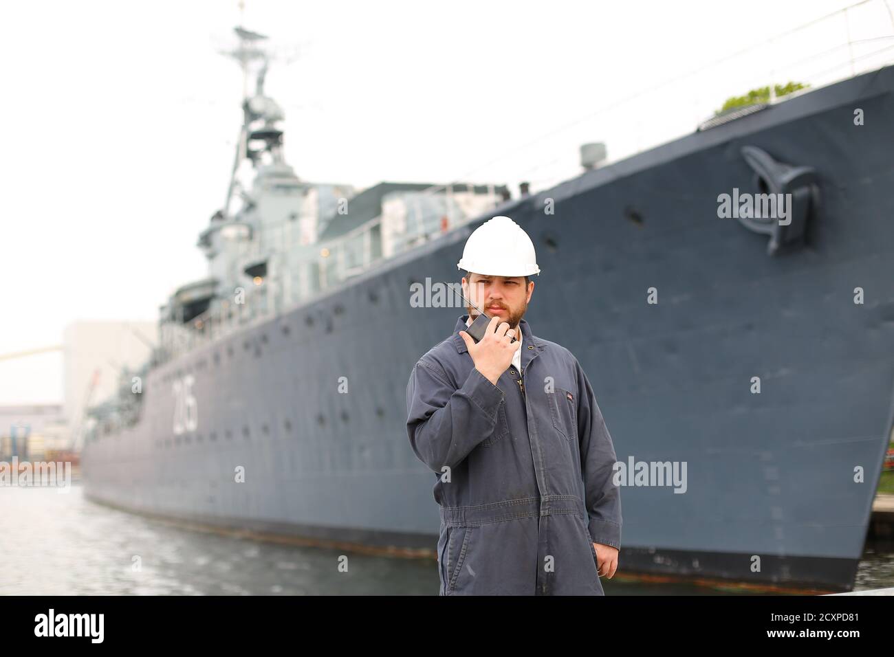 Deck Officer wearing helmet and talking by VHF walkie talkie radio in hands, standing on coast with offshore vessel in background. Concept of maritime Stock Photo