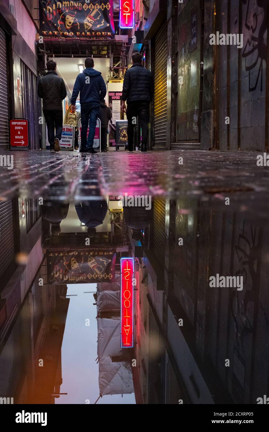 A tattooist neon sign is reflected in a puddle after dusk as pedestrians walk past. The Soho area of London’s West End on a cold and wet January day. Stock Photo