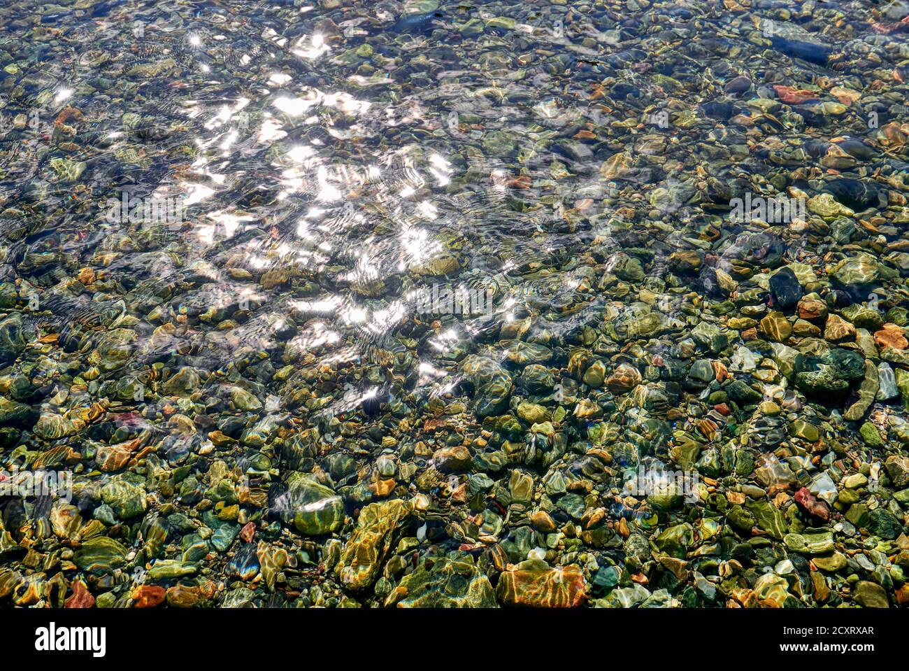 Detail view of a flowing river with sun ray reflections on the water surface, and beautiful colorful pebble stones underwater Stock Photo