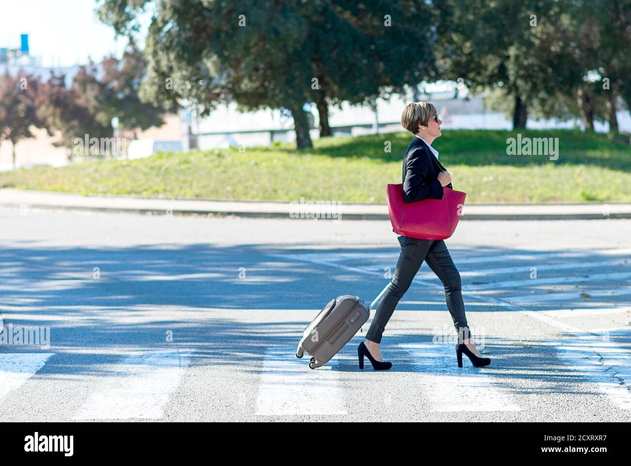Horizontal view of the cheerful business woman carrying the luggage while holding a red shoulder bag Stock Photo